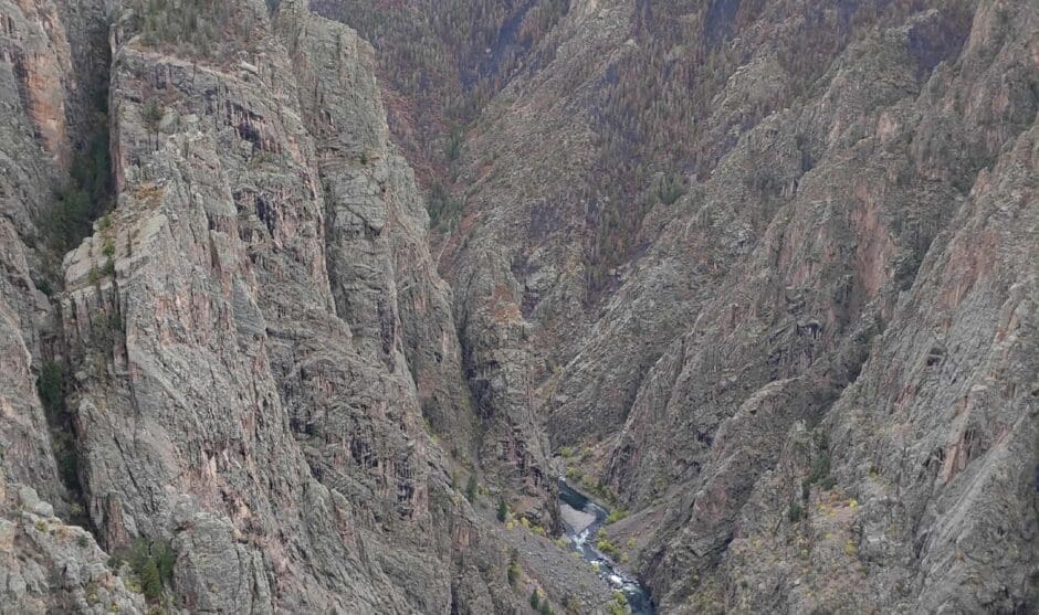 A dramatic, vertical view looking deep into the Black Canyon of the Gunnison. The steep, sheer walls of dark Precambrian rock drop to a winding river far below under an overcast sky.