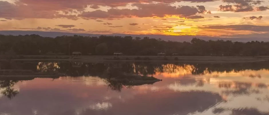 A wide panoramic view of a sunset over a calm body of water at Confluence Park. The sky is filled with soft orange, pink, and purple clouds, which are perfectly mirrored in the still water below. A dark silhouette of a tree-lined shore and small park gazebos sits on the horizon between the sky and the reflection.
