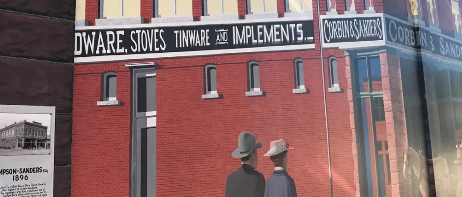 Mural of a red brick historical building with two people in late 19th-century attire viewed from behind. Signs on the building read "Hardware, Stoves, Tinware and Implements" and "Corbin & Sanders." To the left, a plaque shows a historical photo of the Simpson-Sanders Building, dated 1896.