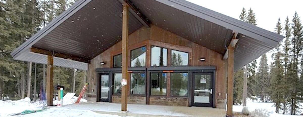 A wide-angle photograph of the newly constructed Skyway Station at the Skyway Trailhead. Visual Details: * Architecture: A modern mountain building with a dramatic, steeply pitched dark grey metal gable roof that extends over a large concrete porch. The exterior features warm wood vertical siding and large glass windows that reflect the sky. * Setting: The building is surrounded by a snowy landscape and a dense forest of evergreen trees under an overcast sky. * Construction: Three sturdy wooden pillars support the roof overhang, and a small orange ladder is visible leaning against the side of the structure, indicating the final stages of the project.