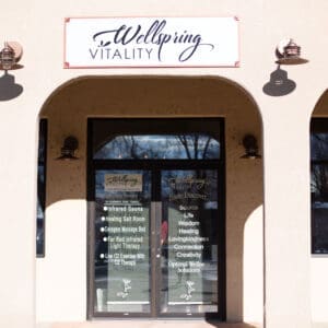 Building Exterior and Entrance
The exterior entrance of Wellspring Vitality, featuring a light-colored stucco building with three large architectural arches. A white rectangular sign above the center arch displays the "Wellspring Vitality" logo in purple script. The glass front doors are adorned with text listing available therapies, including Infrared Sauna, Healing Salt Room, and Red Light Therapy.