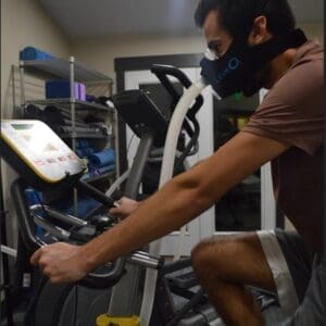 A man is shown from the side practicing "Exercise with Oxygen Therapy" at Wellspring Vitality. He is riding a stationary exercise bike while wearing a large, black LiveO2 respiratory mask connected to a thick, clear oxygen tube. In the background, a fitness rack holds yoga blocks and rolled-up mats, and an elliptical machine is visible.