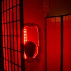 Red Light Therapy Suite
A person seen from behind is seated in a therapy room bathed in deep red light. They have their arms raised in a stretch toward a bright vertical LED red light panel. The space is divided by dark-framed privacy screens with translucent panels, and a small wall hanging above reads "LIGHT" with a Japanese kanji character.