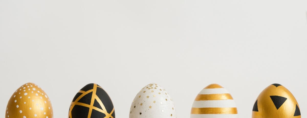 A clean and elegant promotional image for an Easter event. Visual Composition: Main Subject: Five decorative Easter eggs are lined up in a horizontal row against a plain, off-white background. Details: The eggs feature sophisticated modern designs in gold, black, and white, including patterns of dots, geometric lines, small stars, horizontal stripes, and triangles. Style: The lighting is soft and bright, creating a minimalist and festive spring aesthetic.